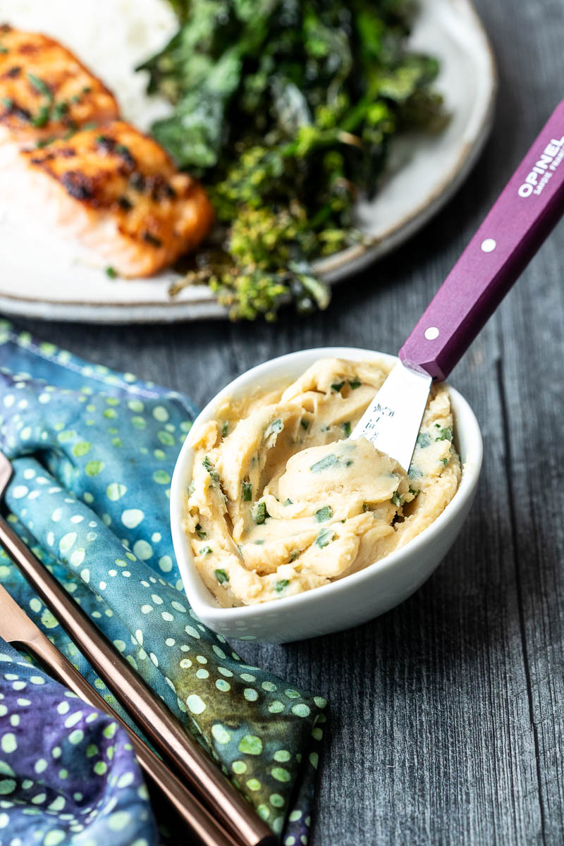 A small bowl of miso butter with herbs next to a spreading knife next to a plate of grilled fish, greens, and rice on a wooden table.