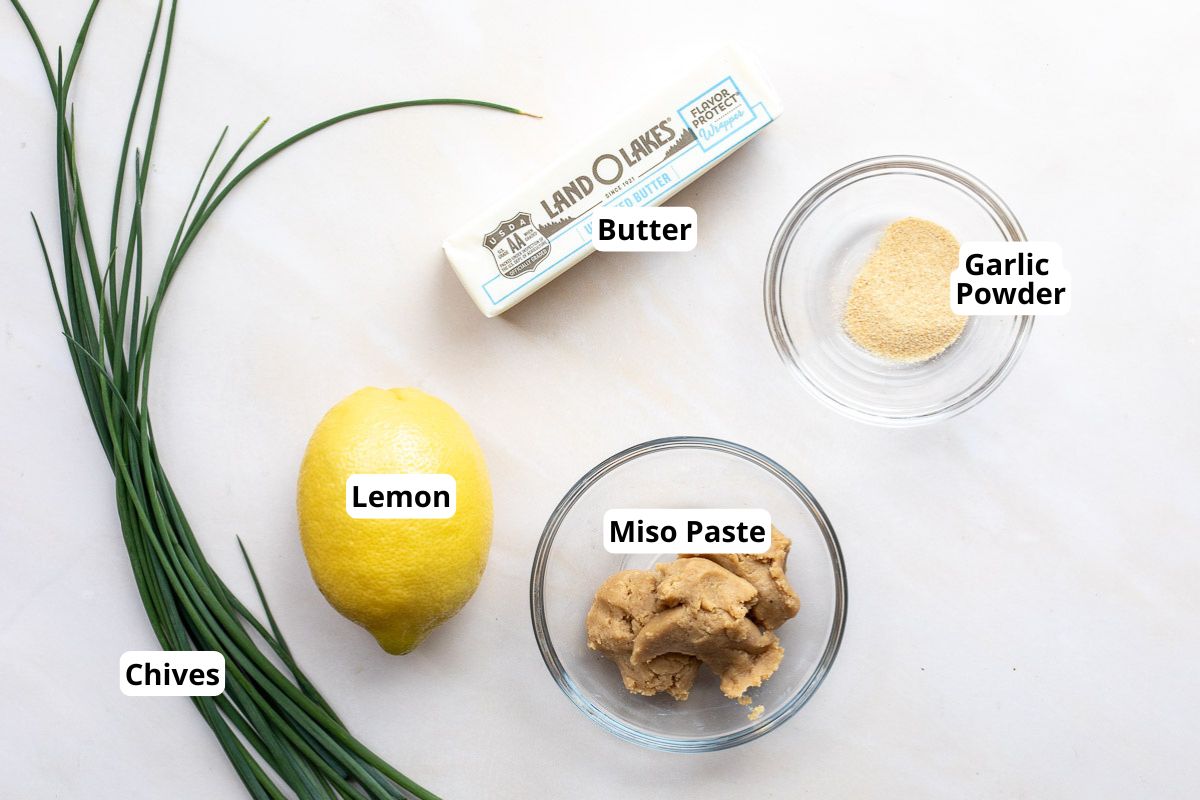 A bundle of chives, a lemon, a stick of butter, a bowl of miso paste, and a bowl of garlic powder are arranged on a white surface, each labeled.