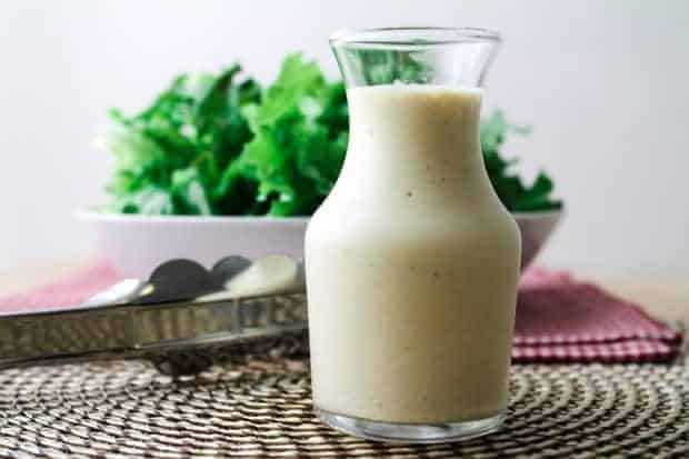 A glass jar filled with a creamy vegan caesar salad dressing sits on a woven placemat, with salad greens and metal tongs in the background.
