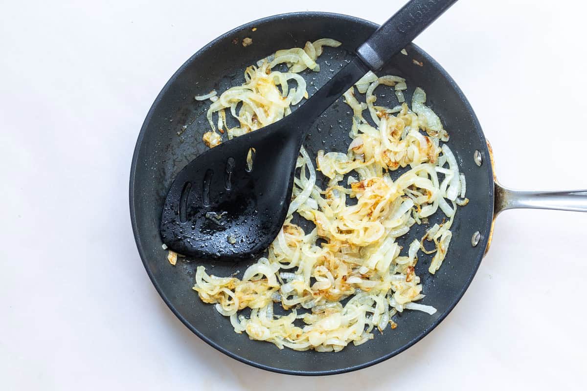 Sliced onions being sautéed and lightly browned in a nonstick frying pan with a black slotted spatula.