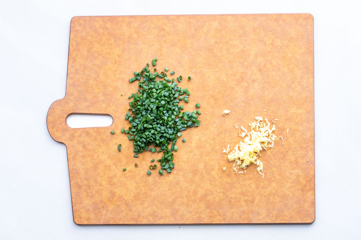 Chopped green chives and grated lemon zest are placed separately on a brown cutting board with a handle.