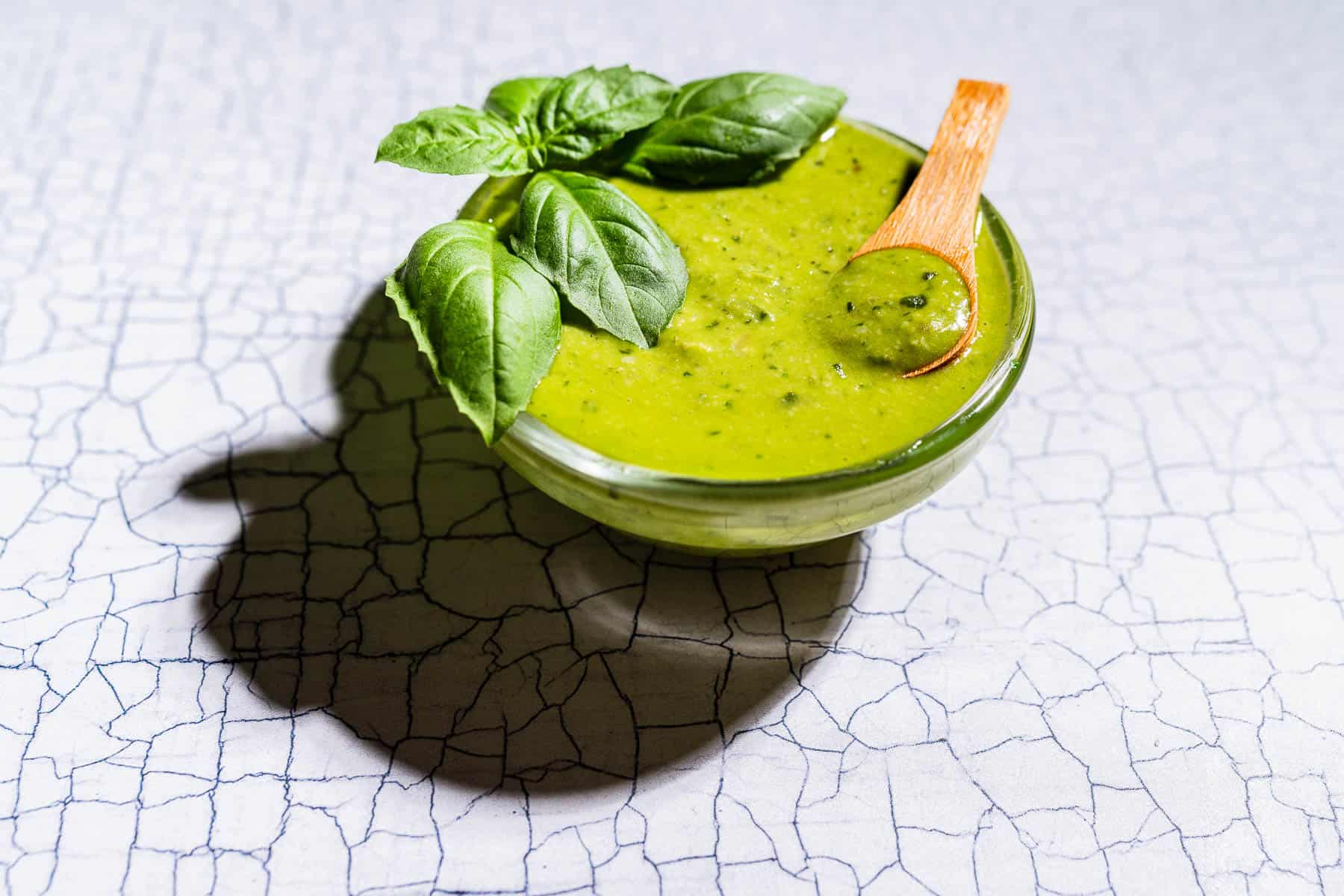 A glass bowl of green pesto salad dressing with basil leaves and a wooden spoon, placed on a textured, cracked white surface.