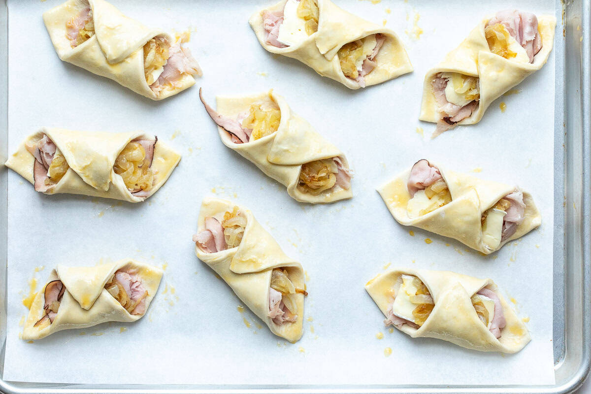 Unbaked puff pastry wraps filled with ham, cheese, and caramelized onions arranged on a parchment-lined baking sheet.