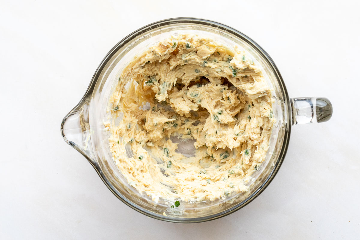 A glass mixing bowl containing a creamy, lightly mixed miso butter with chives on a white surface.