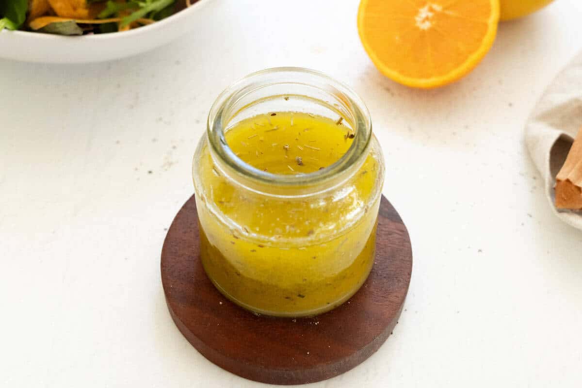 A glass jar of orange vinaigrette sits on a round wooden coaster, with a halved orange and a bowl of salad visible in the background.