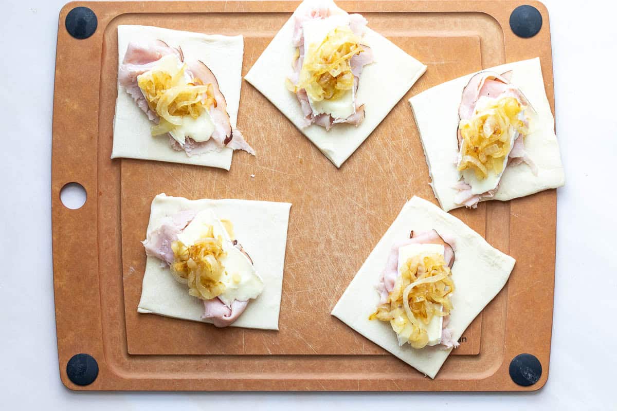 Five pieces of puff pastry dough on a cutting board topped with ham, cheese, and caramelized onions, ready to be folded or baked.