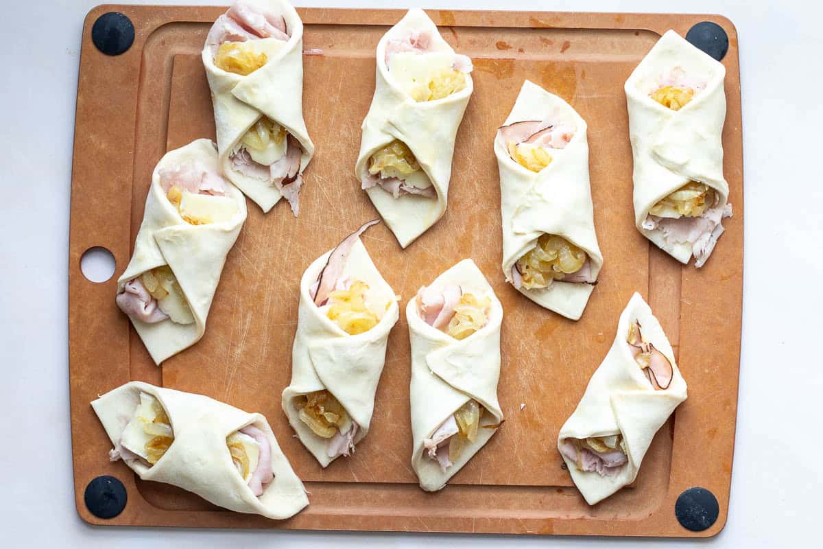 Nine unbaked puff pastry turnovers filled with ham, cheese, and caramelized onions are arranged on a brown cutting board.