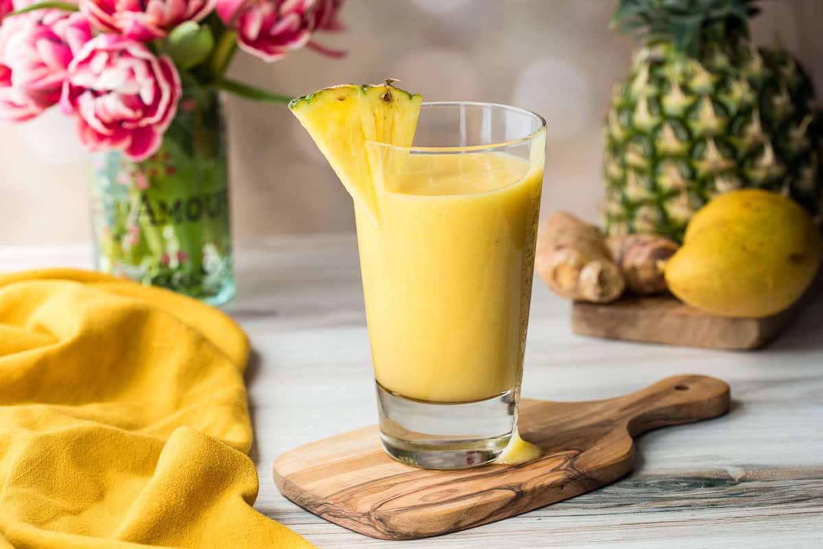 A glass of pineapple mango smoothie garnished with a pineapple slice sits on a wooden board, surrounded by a pineapple, mango, ginger, and pink flowers.