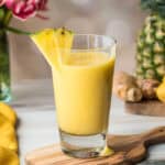A glass of yellow mango pineapple smoothie garnished with a pineapple wedge, placed on a wooden board with pineapple, ginger, and flowers in the background.