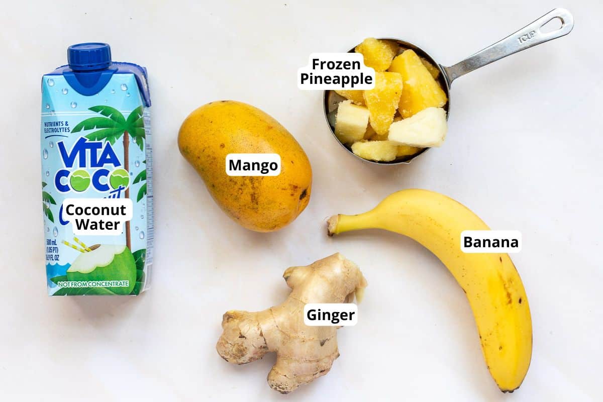 Carton of coconut water, a whole mango, a measuring cup with frozen pineapple, a banana, and a piece of ginger on a white surface, each labeled.