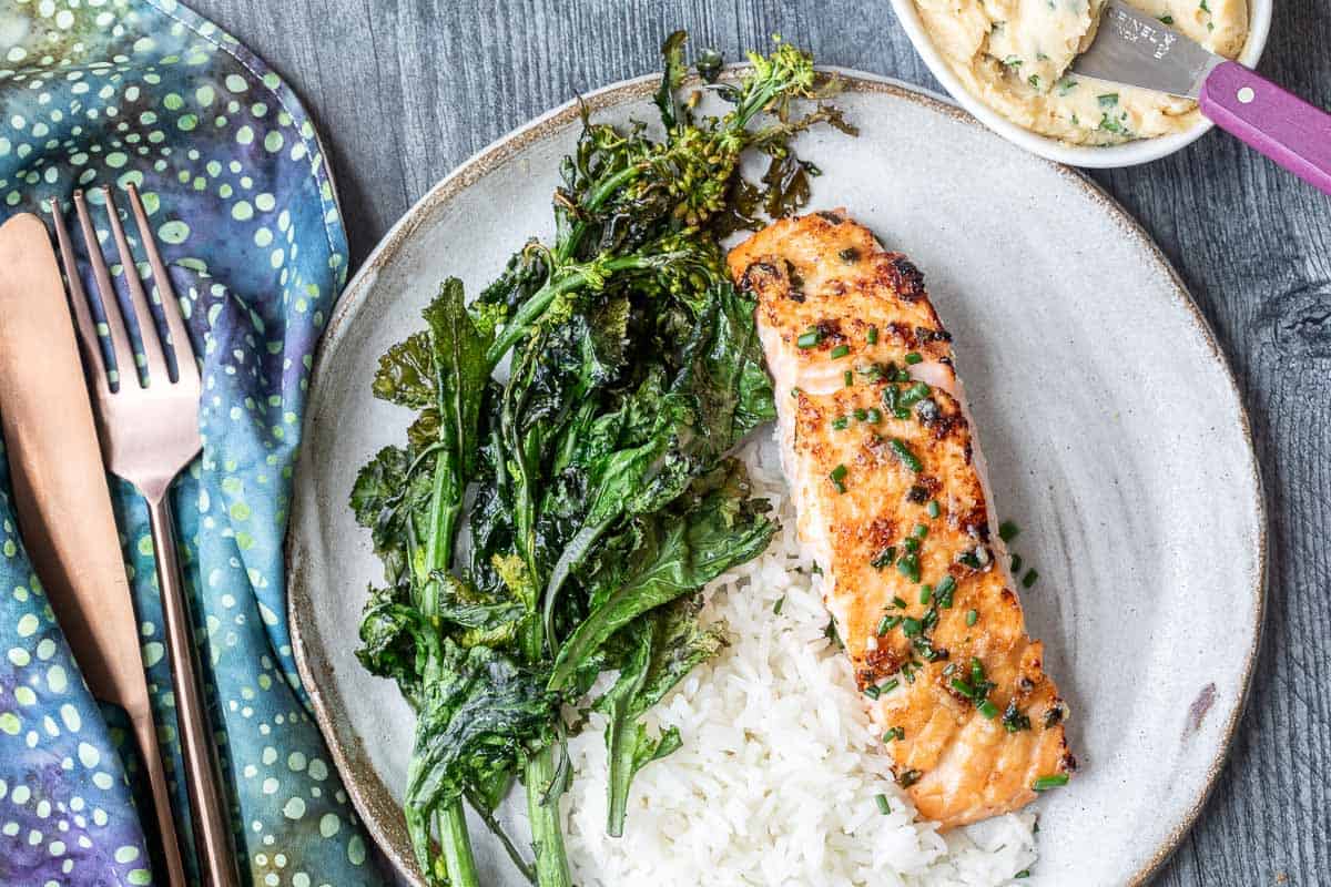 A plate of miso salmon on white rice with sautéed greens, next to a napkin, utensils, and a dish of miso butter on a dark wood table.