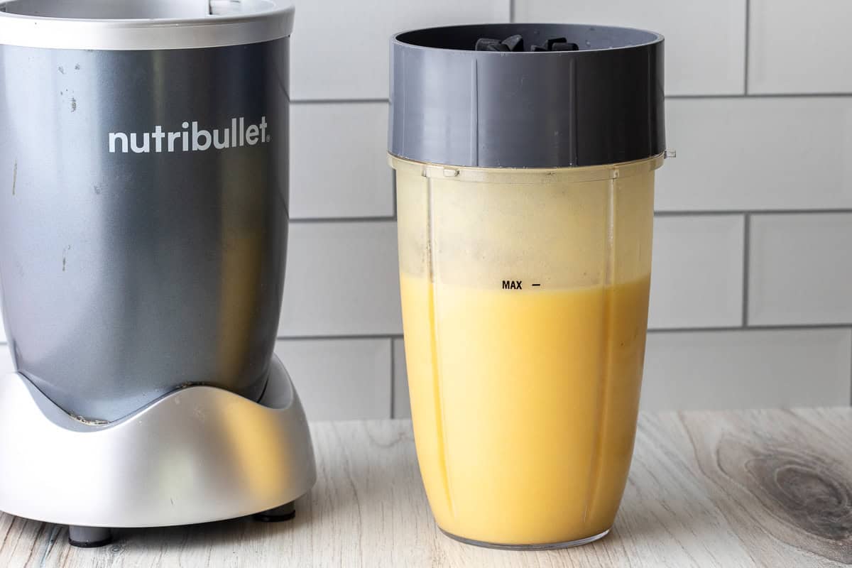 A Nutribullet blender base sits next to a blending cup filled with yellow mango pineapple smoothie, placed on a wooden surface in front of a white tiled wall.