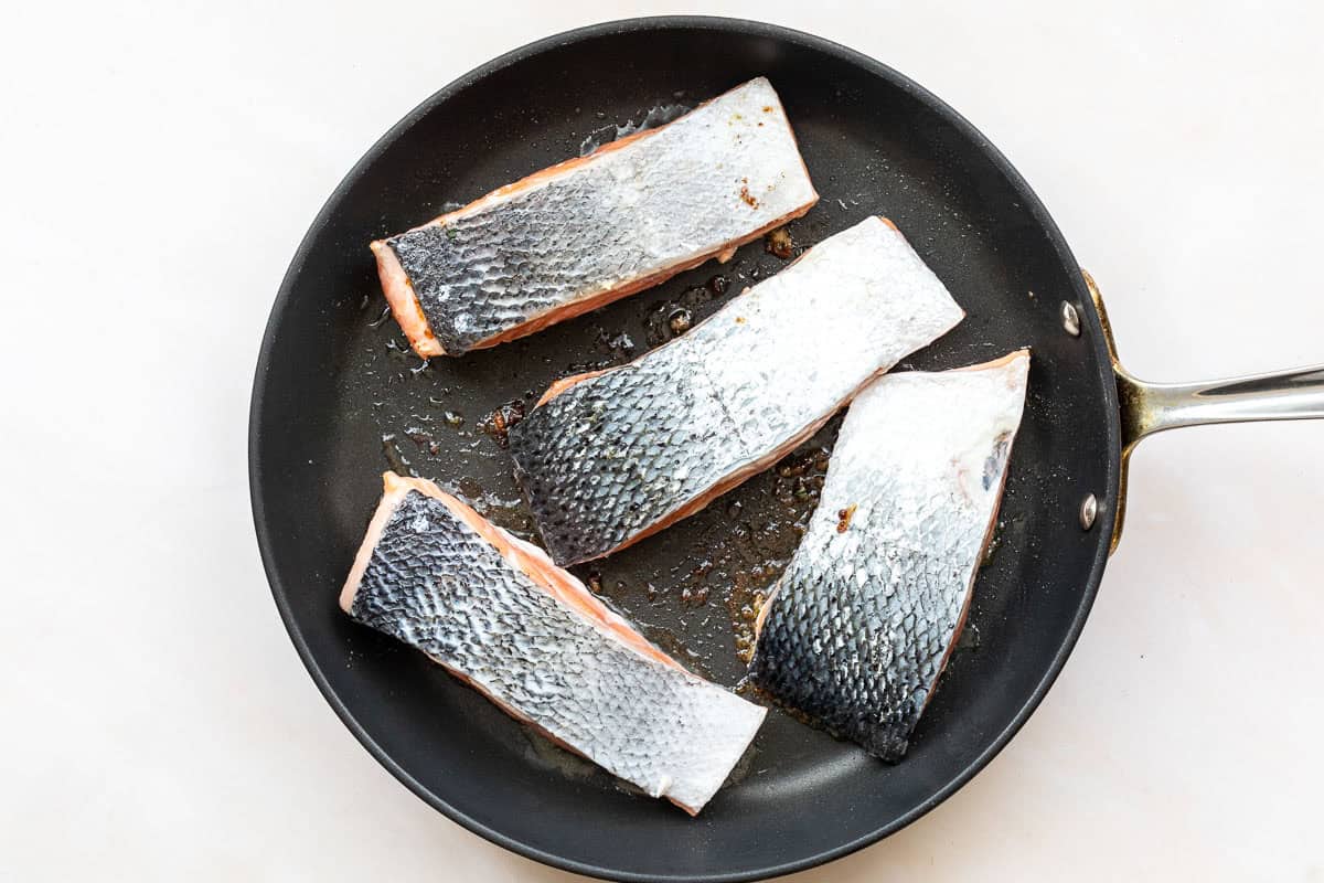 Three raw salmon fillets with skin on are placed skin side down in a black frying pan on a white surface.