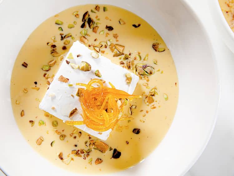 Ile Flottante sits in a creamy yellow sauce, topped with orange zest and chopped nuts, served in a white bowl.