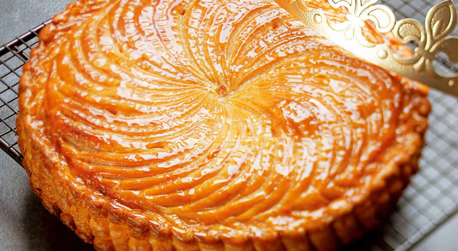 A golden, glossy Galette des Rois pastry with detailed patterns sits on a cooling rack beside a decorative gold paper crown.