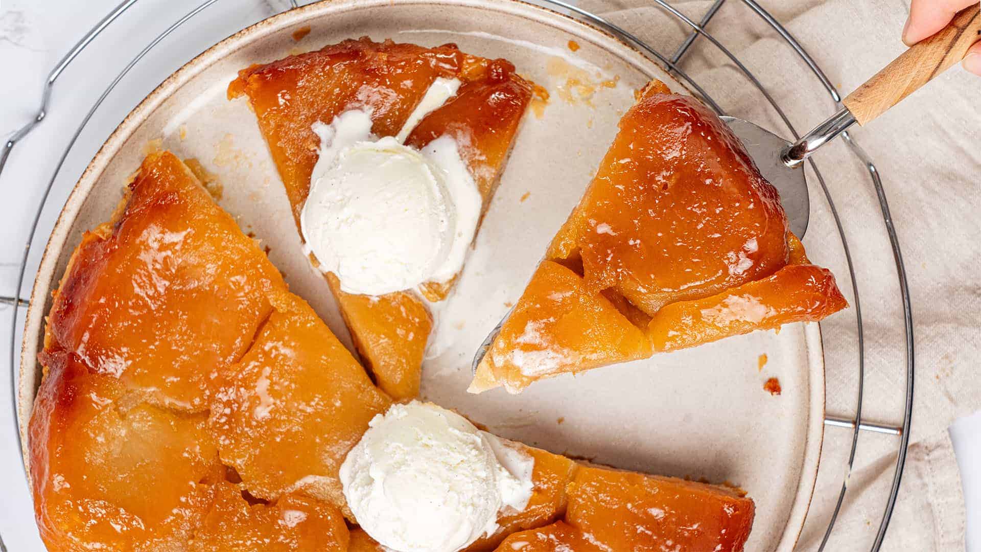 A sliced caramelized apple Tarte Tatin sits on a plate, topped with two scoops of vanilla ice cream; one slice is being lifted with a spatula.