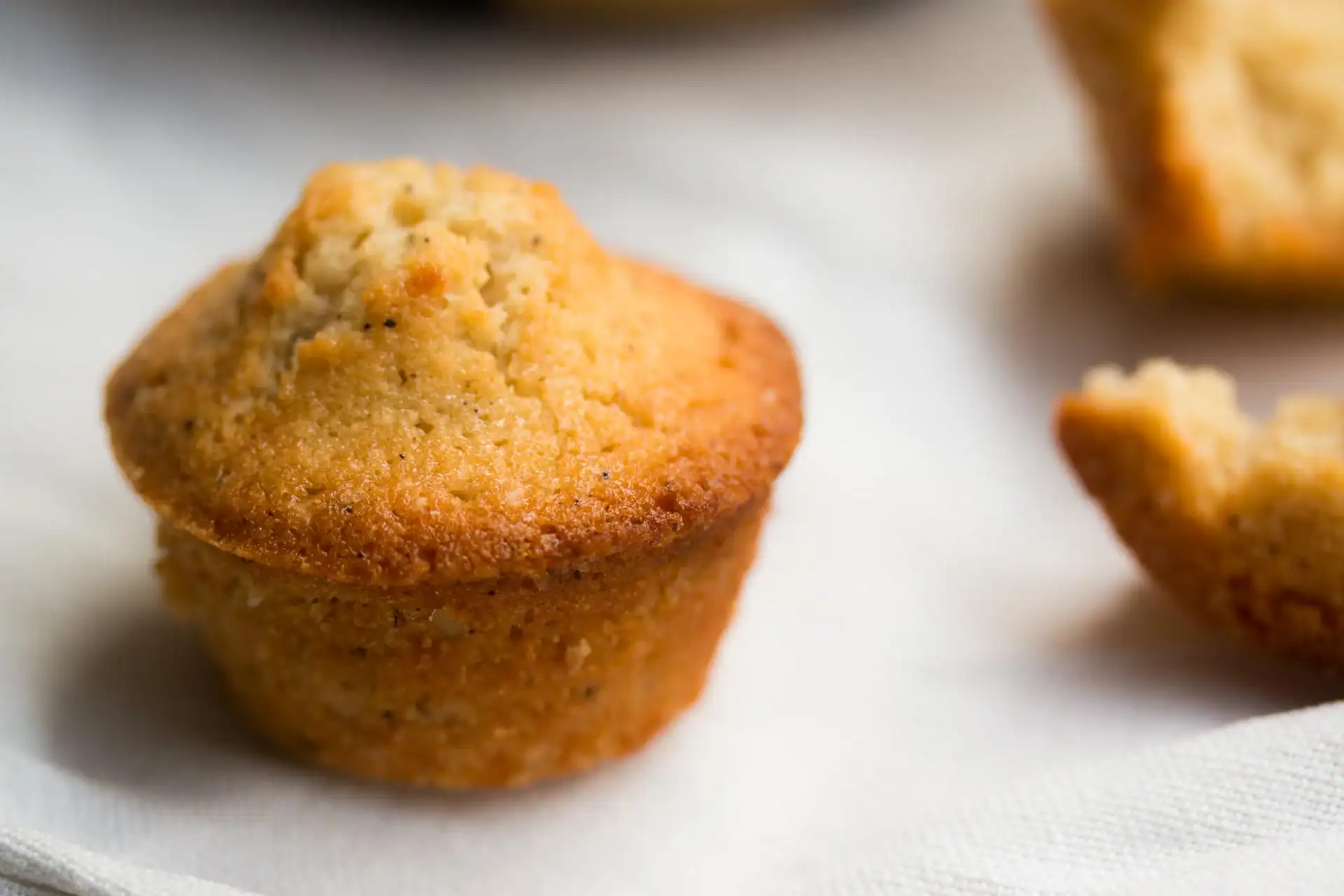 A close-up of a golden brown financier with a domed top, resting on a white cloth next to a partially visible, broken financier.