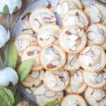 A plate of almond financier cakes topped with sliced almonds, placed next to white artificial flowers and green leaves.