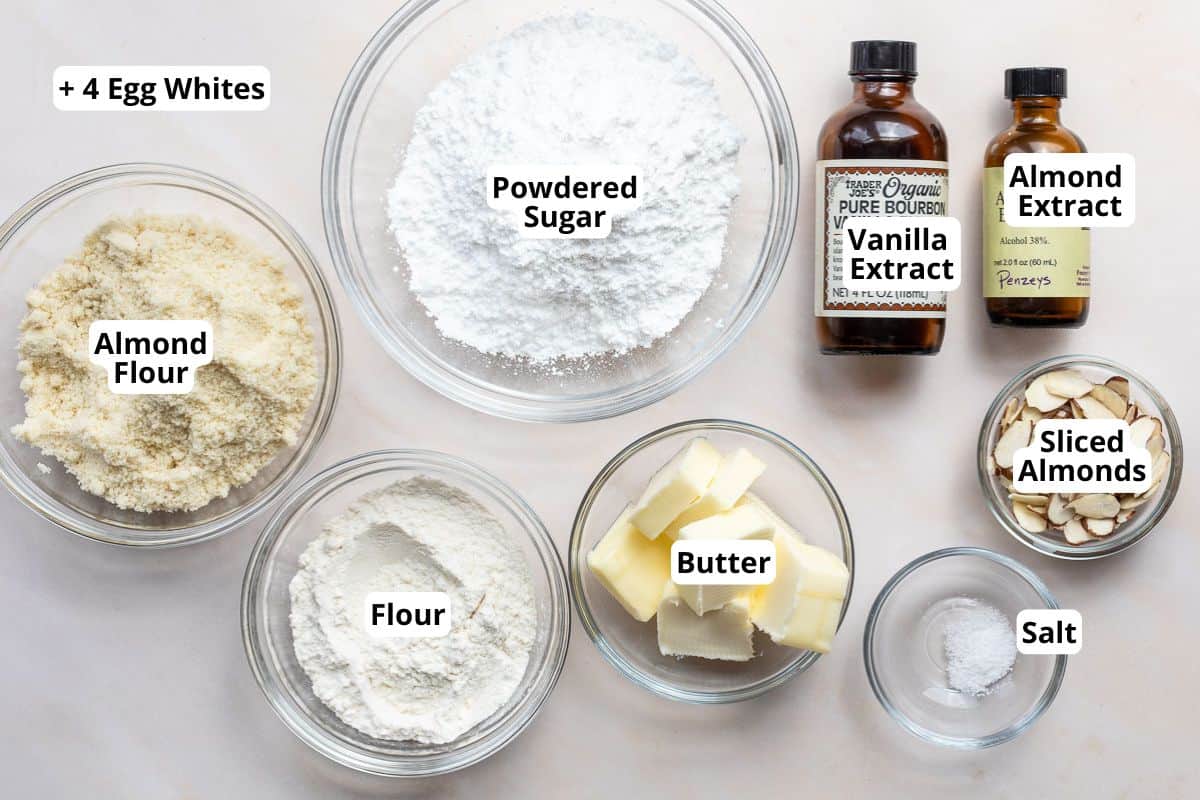 Baking ingredients including egg whites, powdered sugar, almond extract, vanilla extract, almond flour, flour, butter, sliced almonds, and salt are displayed in labeled bowls and bottles.