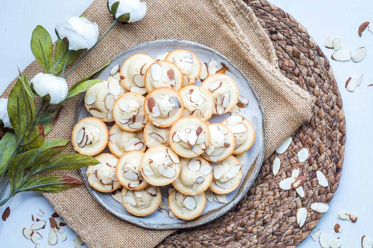 A plate of almond financiers topped with sliced almonds sits on a burlap mat, with white flowers and almond slices scattered nearby.