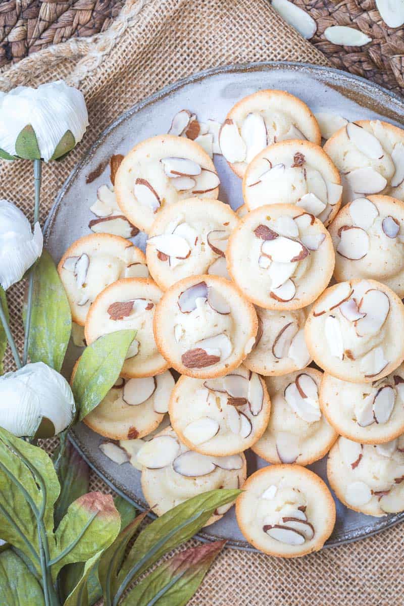 A plate of almond financiers topped with sliced almonds sits on burlap next to artificial white flowers and green leaves.