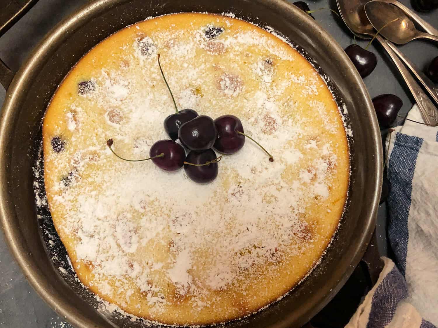 A round baked clafoutis dusted with powdered sugar, topped with fresh cherries, in a metal baking pan next to a napkin and some spoons.