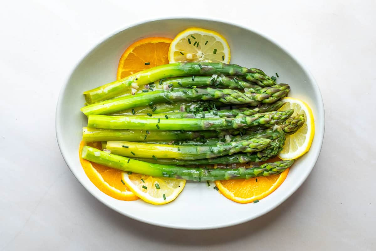 A plate of steamed asparagus spears drizzled with citrus vinaigrette, garnished with chopped chives and served with orange and lemon slices.