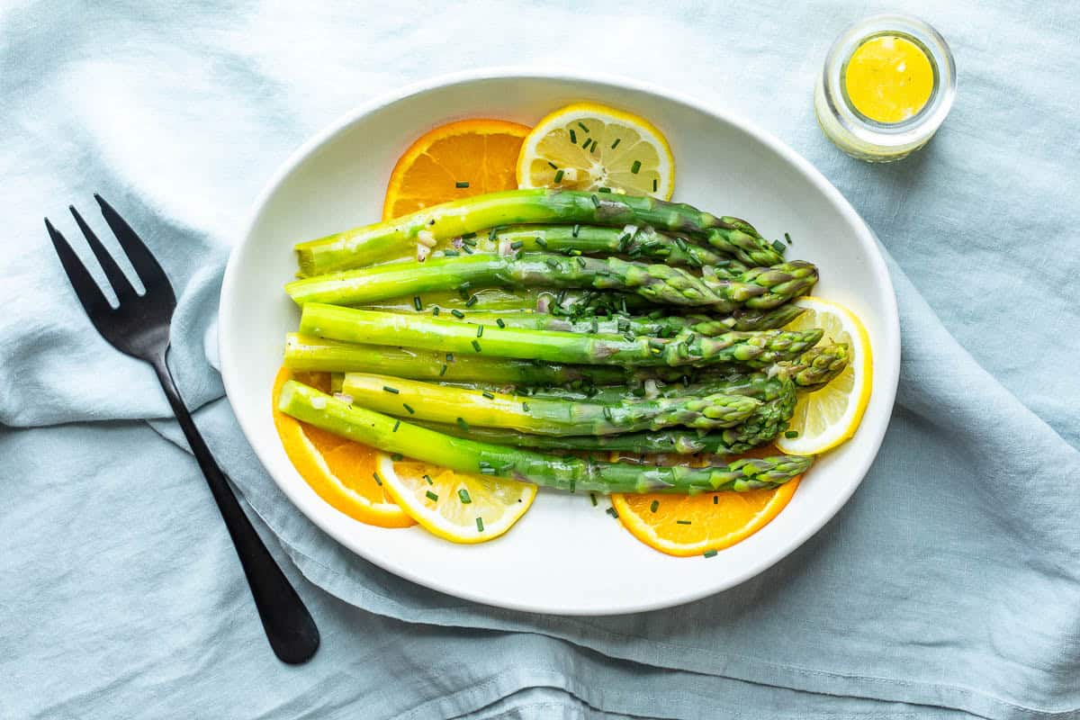Steamed asparagus spears served on a white plate with lemon and orange slices, garnished with chopped chives, next to a small jar of citrus vinaigrette.