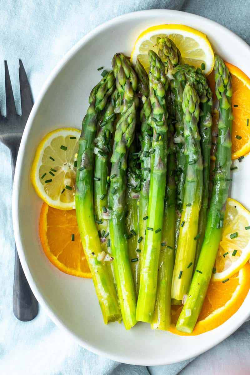 Steamed asparagus spears served on a white plate with orange and lemon slices, drizzled with citrus vinaigrette and garnished with chopped chives. A fork is placed beside the plate.