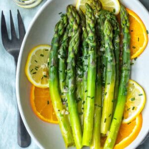 Steamed asparagus spears drizzled with citrus vinaigrette and garnished with chopped chives, served on a white plate with orange slices. A fork and knife are placed beside the plate.