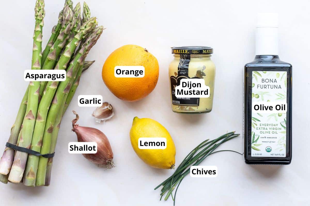 A bundle of asparagus, an orange, a jar of Dijon mustard, a bottle of olive oil, a bulb of garlic, two shallots, a lemon, and several chives are arranged on a white surface.