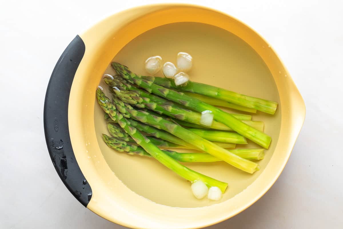 Several stalks of asparagus are submerged in a bowl of ice water with a few ice cubes floating on the surface.
