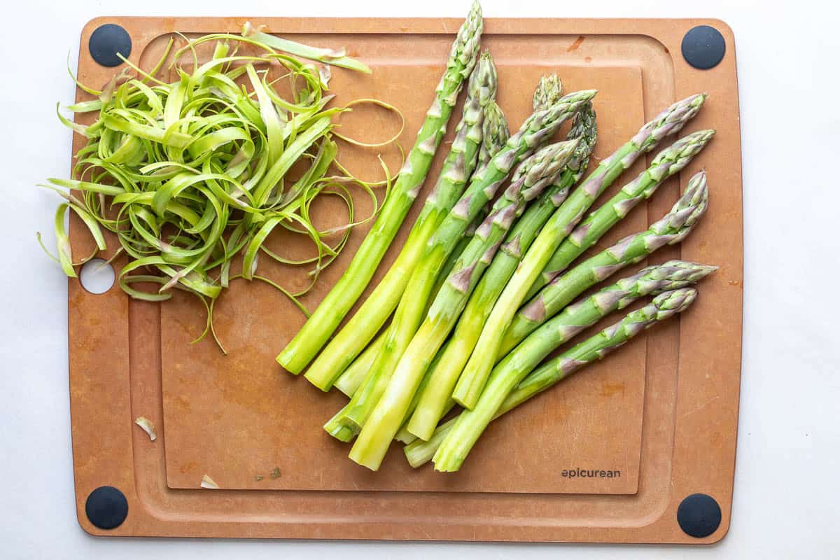 A bunch of peeled asparagus spears and a pile of asparagus peelings on a brown cutting board.
