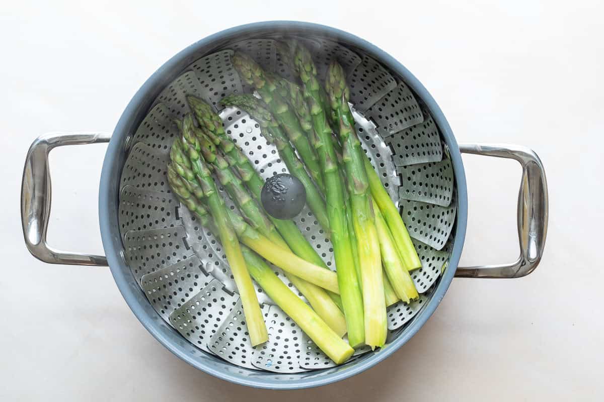 Several stalks of asparagus are being steamed in a metal steamer basket inside a pot.