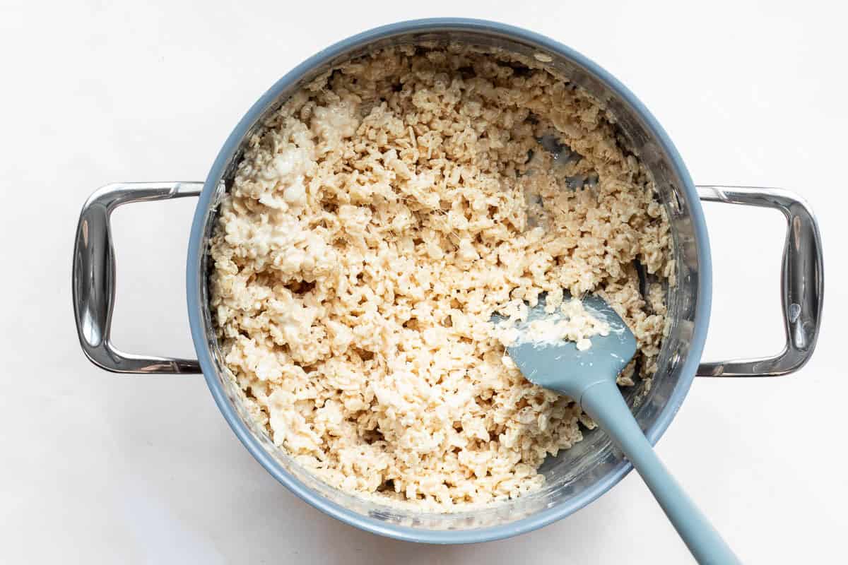 A metal pot filled with a mixture of Rice Krispies, browned butter, and melted marshmallows, with a blue spatula resting inside.