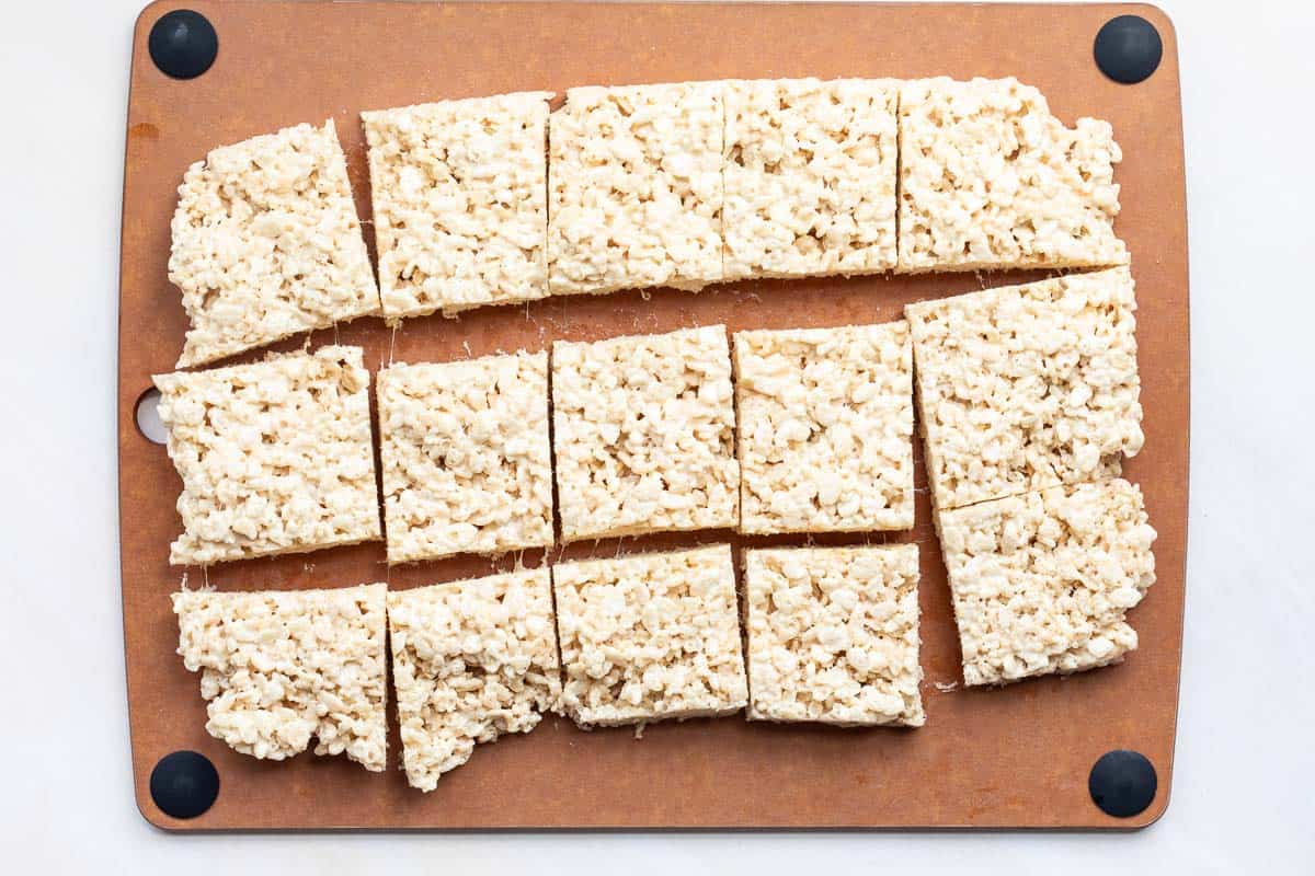 Fifteen Brown Butter Rice Krispies treats cut into squares are arranged on a brown cutting board with black corner grips.
