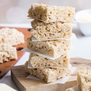 A stack of brown butter rice krispie treats separated by parchment paper sits on a wooden board, with more treats visible in the background.