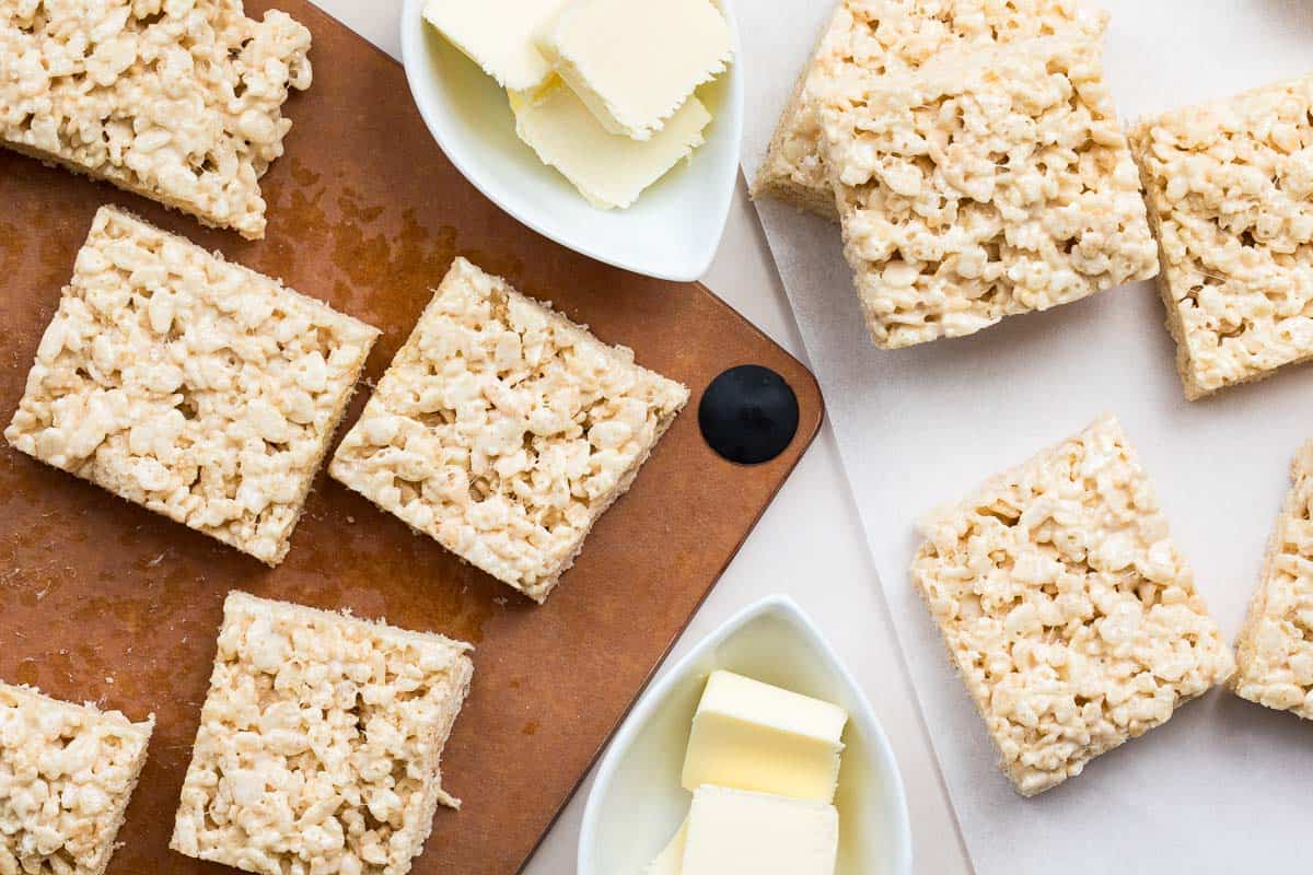 Browned Butter Rice Krispie treats cut into squares are placed on a brown cutting board and white surface, with small dishes of sliced butter nearby.