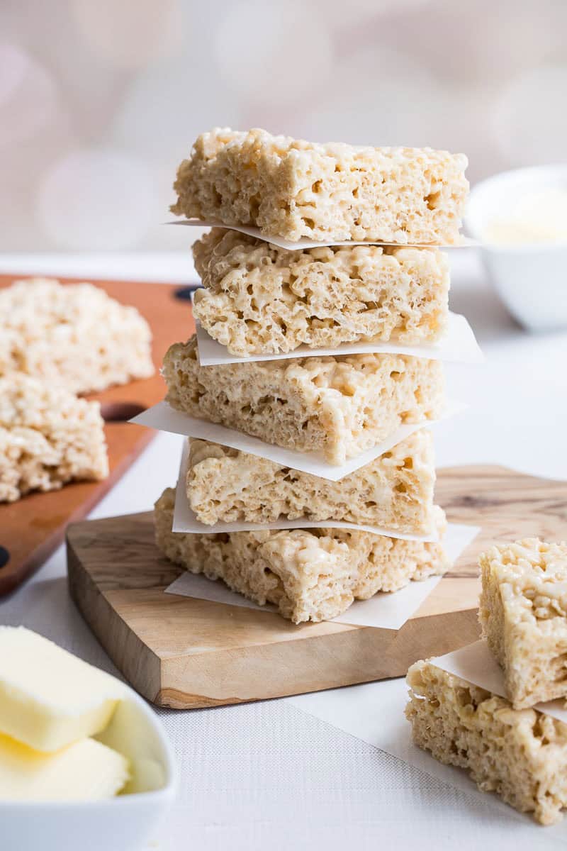A stack of five brown butter Rice Krispies treats separated by parchment paper sits on a wooden board; more treats and a bowl of butter are visible in the background.