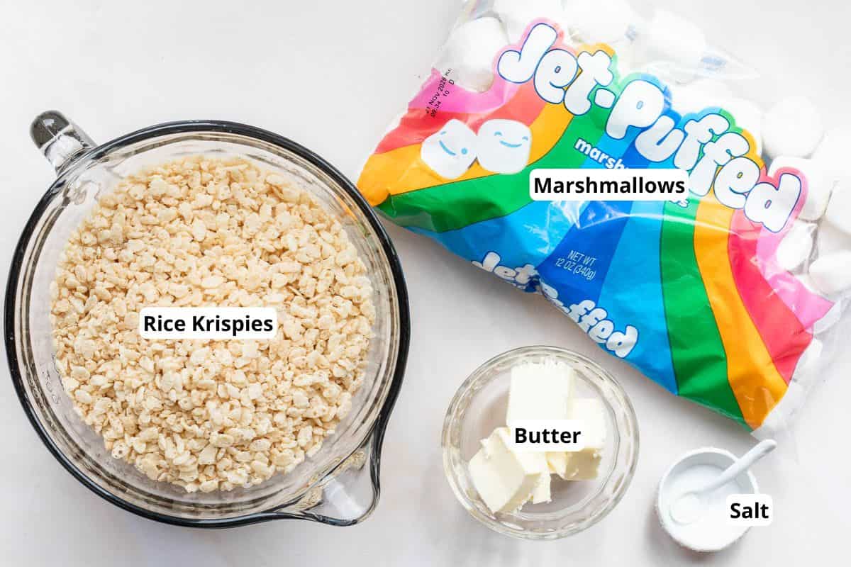 A bowl of Rice Krispies, a bag of Jet-Puffed marshmallows, a dish of butter, and a small bowl of salt are arranged on a white surface.