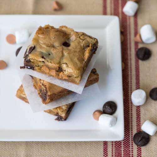 Three blondies stacked on a white plate, with chocolate chips, butterscotch chips, and mini marshmallows scattered around on a tan and red striped cloth.