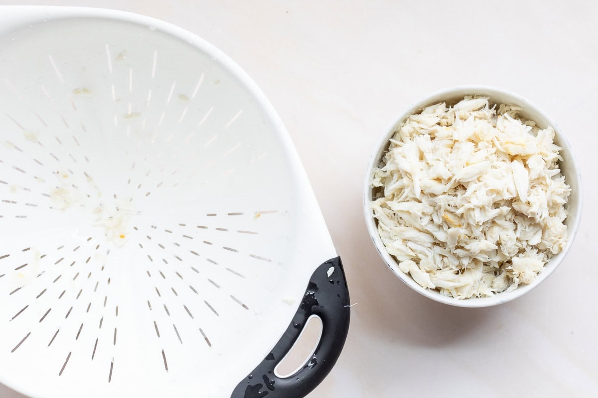 A white colander sits next to a bowl filled with shredded crab meat on a light surface.