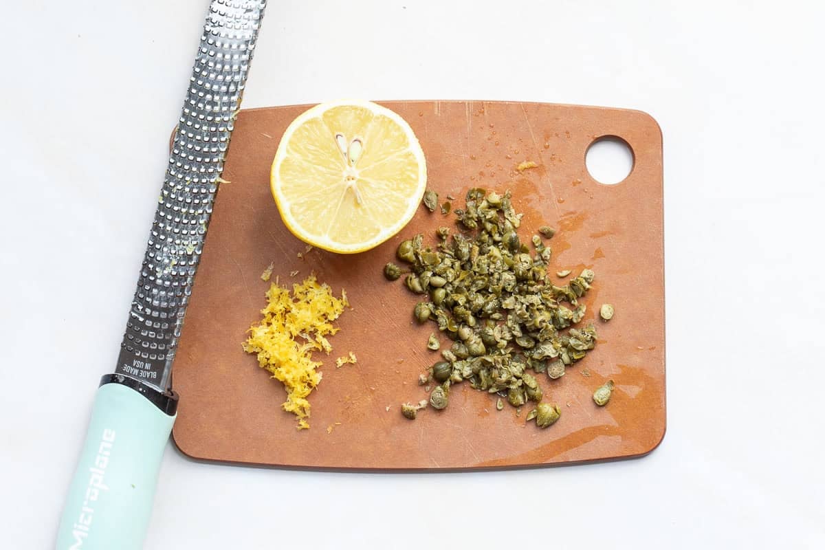 A grater, half a lemon, lemon zest, and chopped capers are arranged on a small brown cutting board.