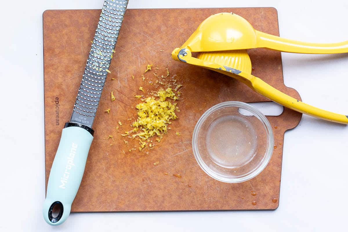 Microplane grater, lemon squeezer, lemon zest, and a small bowl of lemon juice on a brown cutting board.