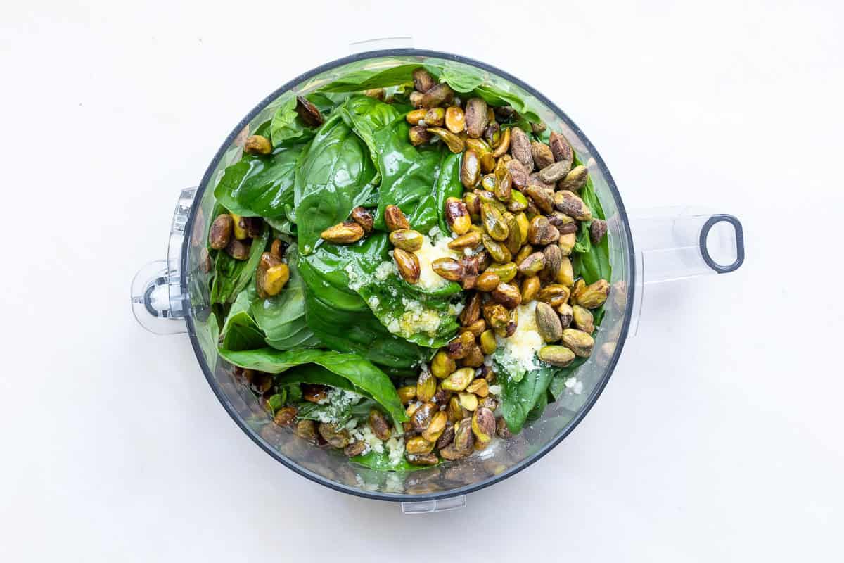 Overhead view of a food processor bowl filled with basil leaves, pistachios, grated cheese, and other pesto ingredients on a white background.