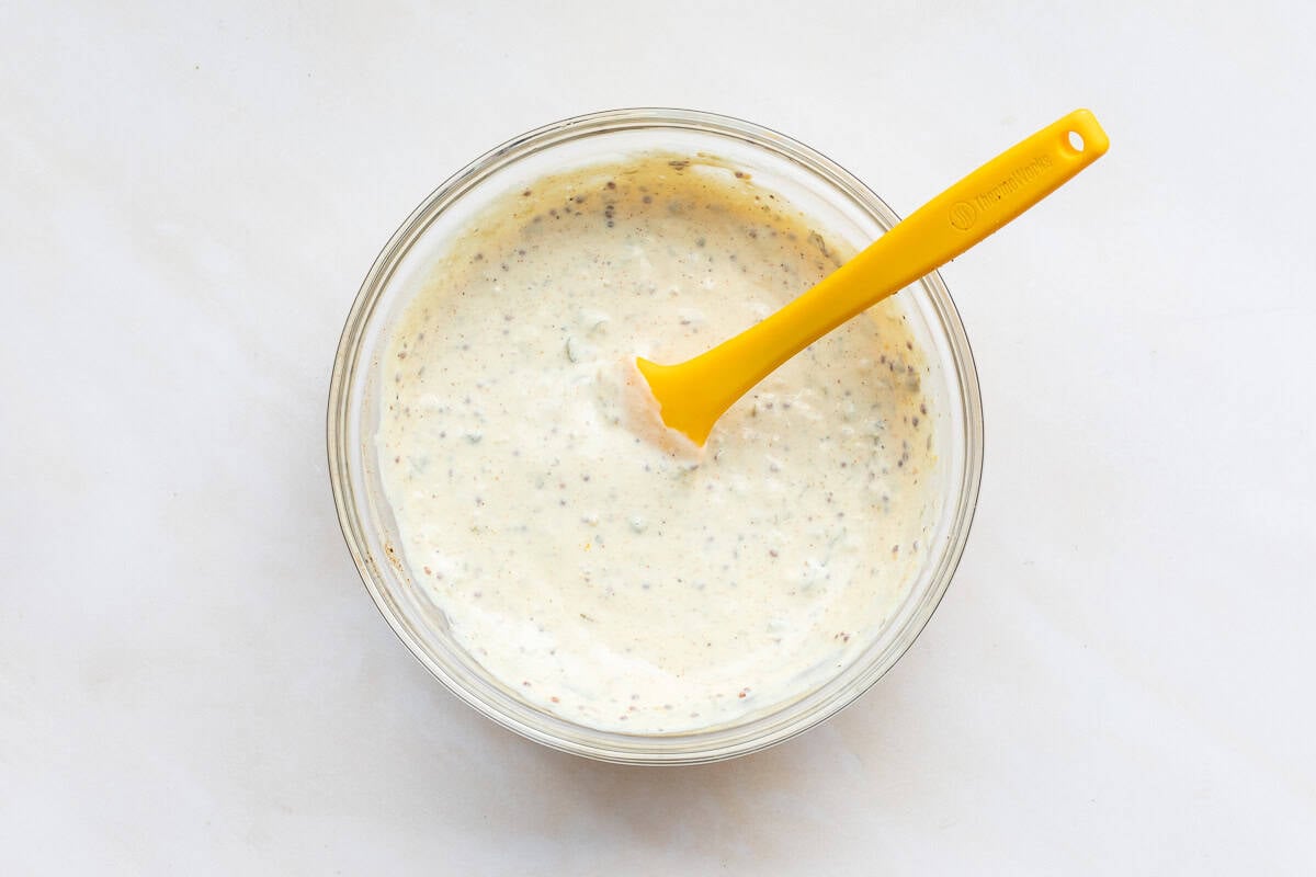 A glass bowl filled with creamy sauce for crab cakes speckled with herbs, stirred with a yellow silicone spatula, sits on a white surface.