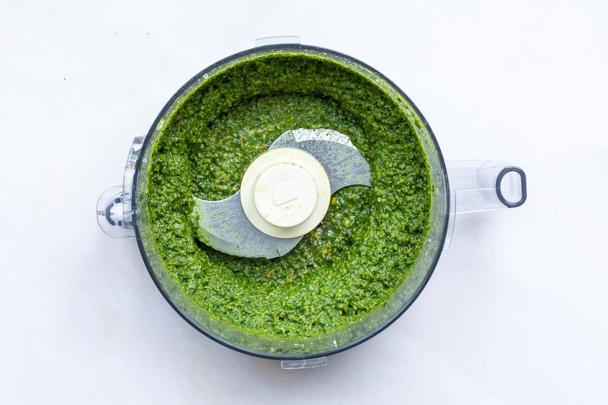 Overhead view of pistachio pesto sauce in a food processor with a silver and white blade, against a white background.