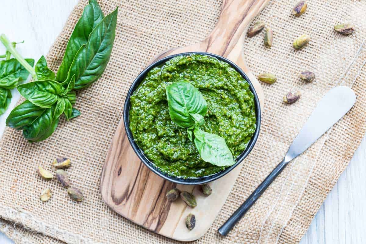 A bowl of pistachio pesto garnished with basil leaves, with a spreading knife and scattered pistachios on a wooden board and burlap cloth.