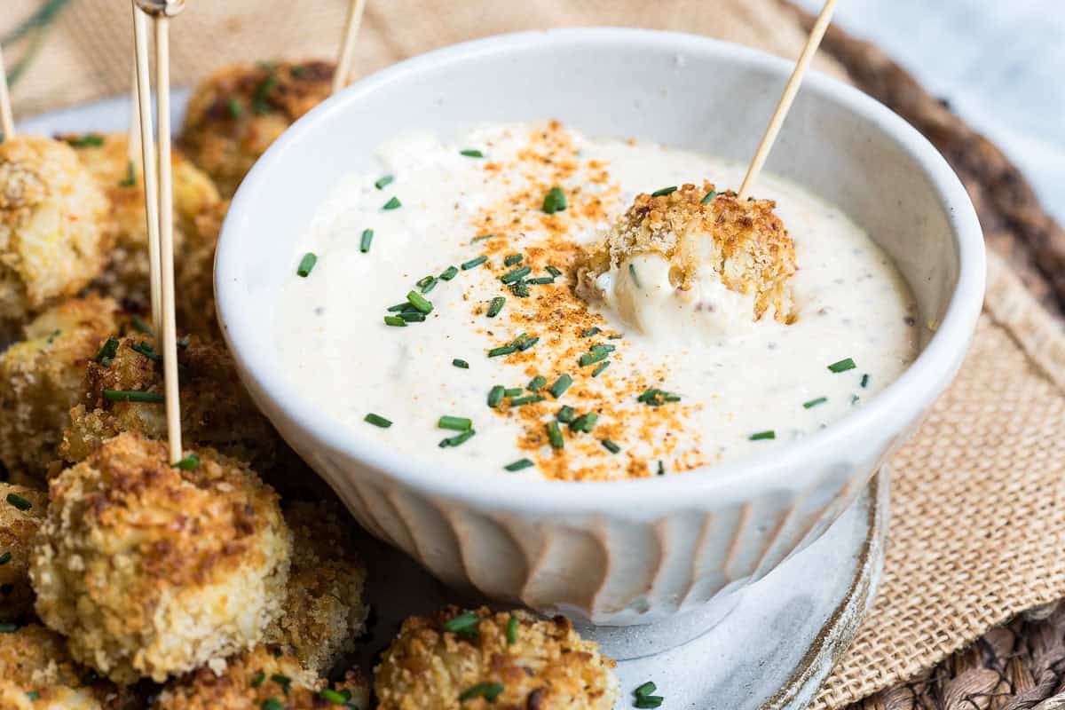 Breaded crab balls on skewers are served with a bowl of creamy dipping sauce, garnished with chopped chives and seasoning. One crab ball is dipped in the sauce.