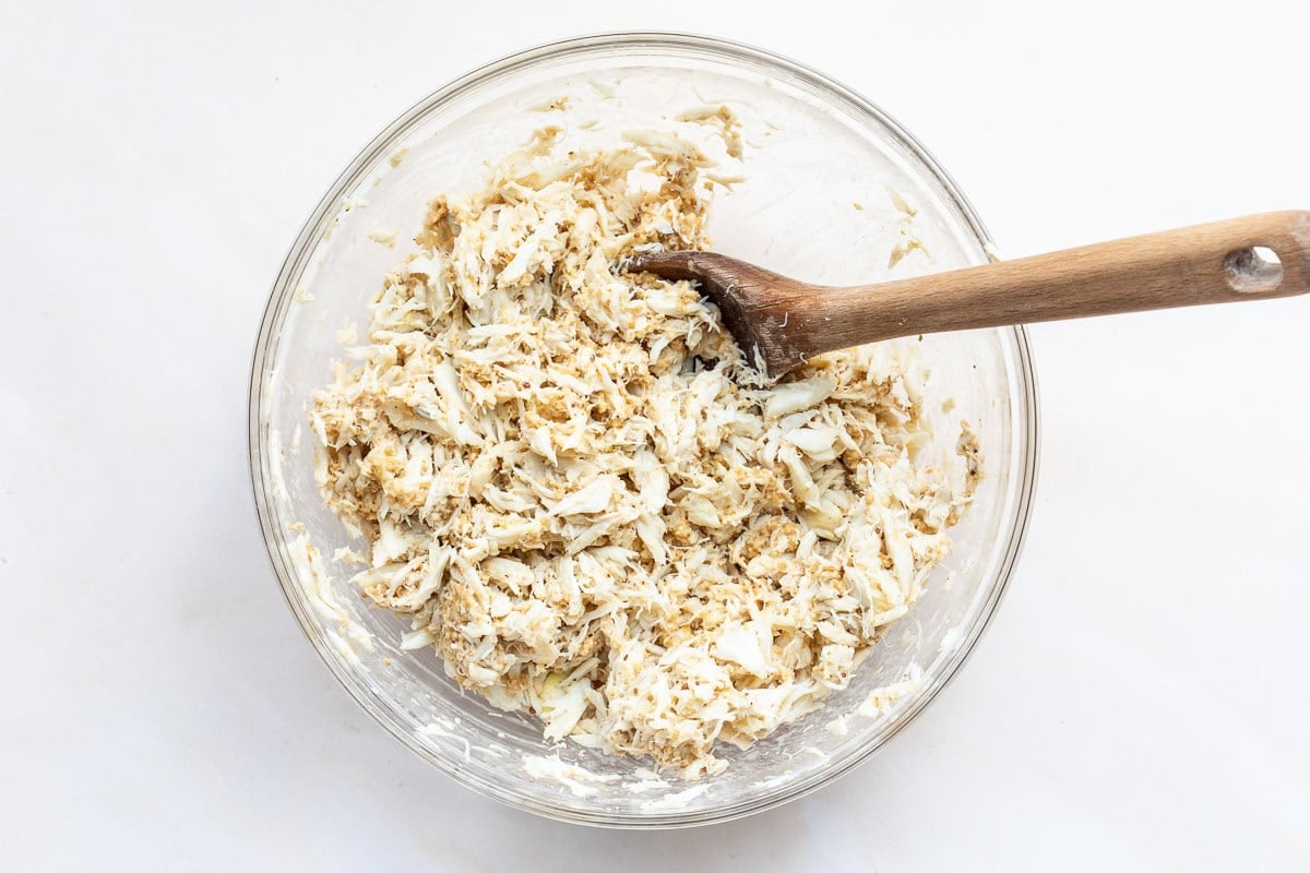 A clear glass bowl filled with a mixture of shredded crab meat and breadcrumbs, with a wooden spoon resting inside.